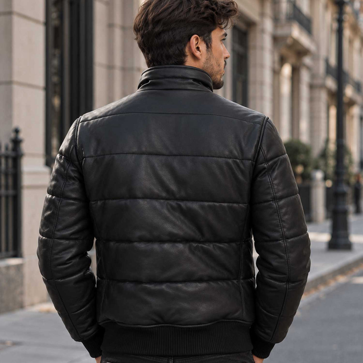 A man viewed from behind wearing a black leather puffer jacket with horizontal quilting, standing on a city street.