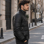 A man wearing a black quilted  puffer jacket stands in profile on a city street.